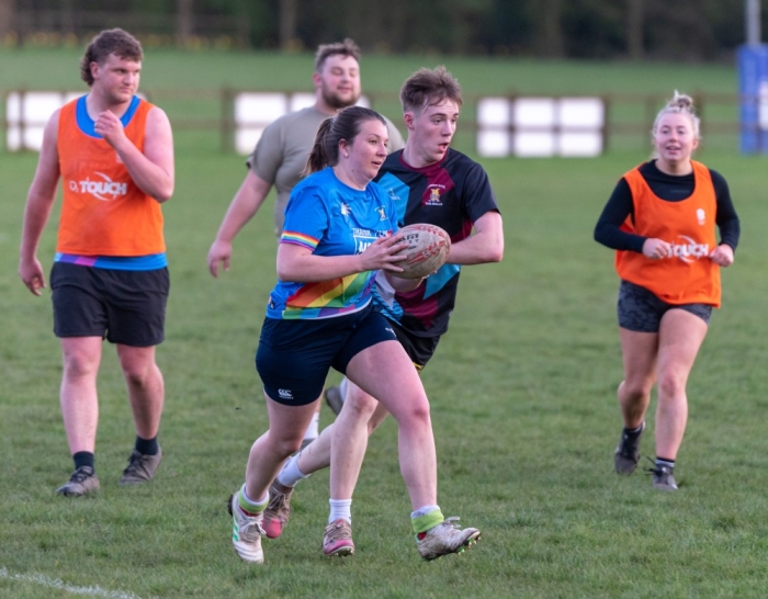 A rugby player is pursued by an opponent as she runs with the ball during a mixed-sex game, with other players looking on in the background.