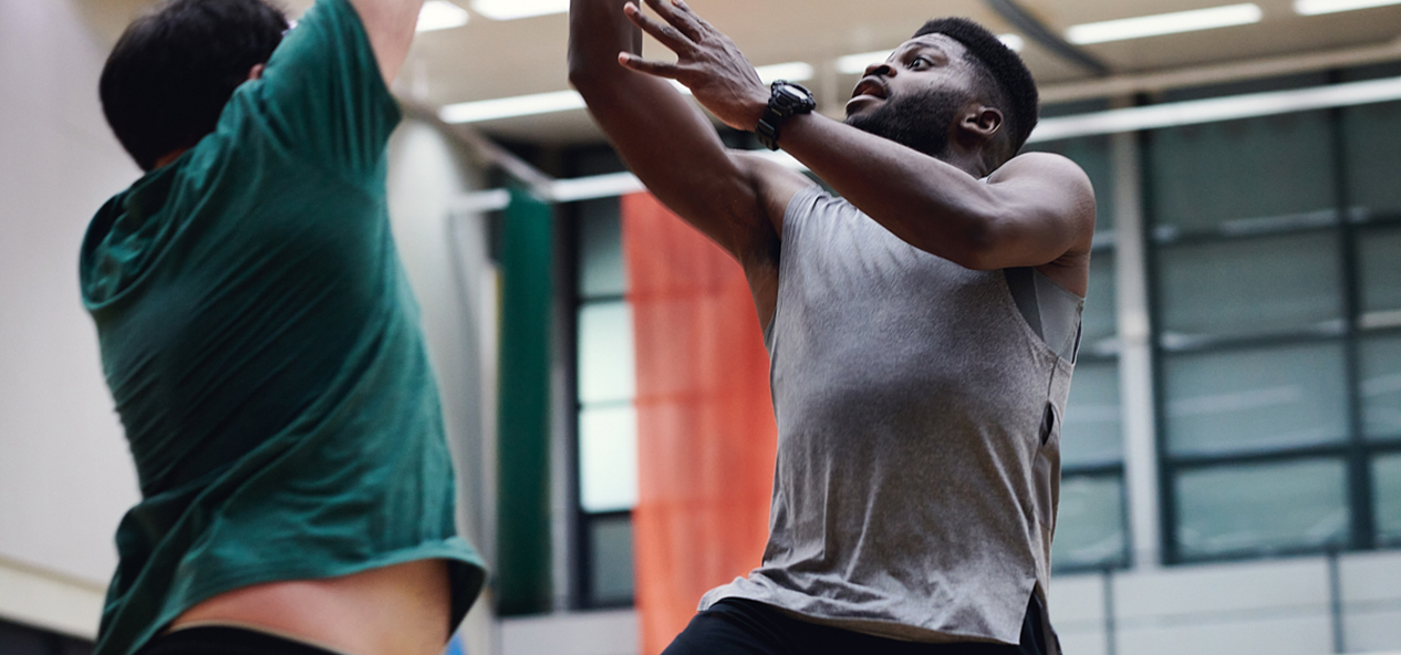 Two men jump while playing basketball on and indoors court.