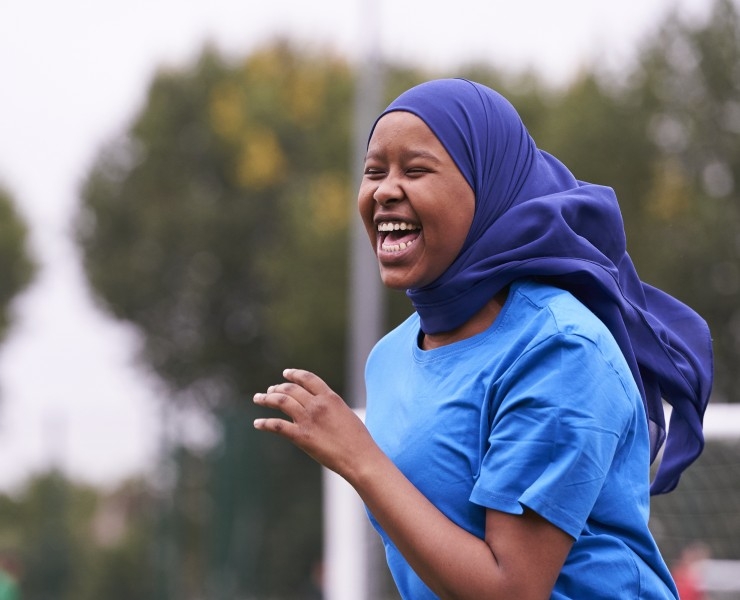 A girl wearing a headscarf laughs on an outdoor sports pitch.