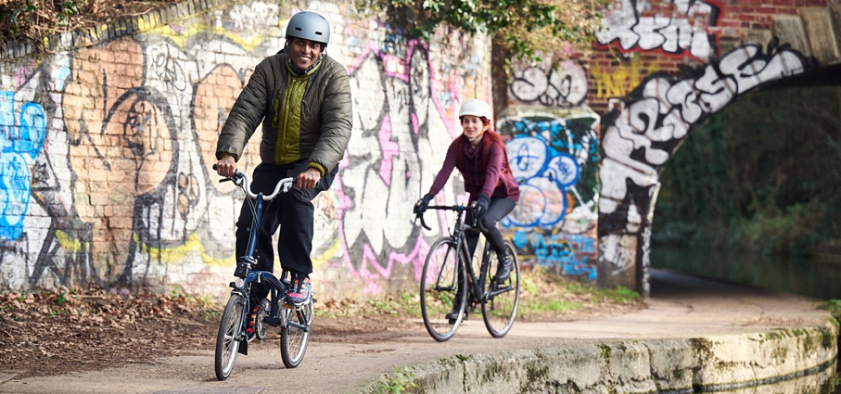 Couple wearing helmets cycle down the side of a canal in line