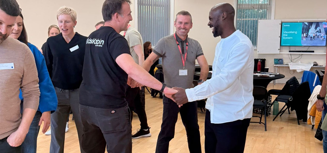 Three men smile and hold each other's hands as they go in circles in a room with a Leading the Movement presentation showing on a screen in the background. 